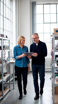 Two engineers reviewing tablet while walking through bright electronics laboratory