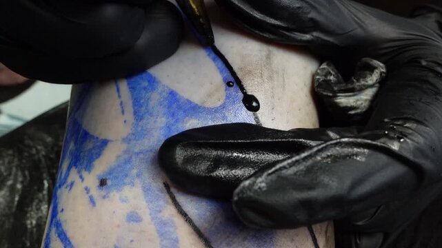 Close-up of a tattoo artist in black gloves drawing a fresh black ink outline over a blue transfer stencil on skin