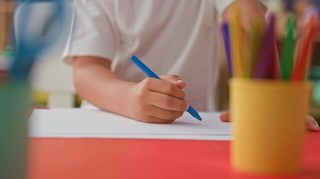 Boy drawing with blue crayon, hand on paper, sketching a face near a yellow pencil holder on red table in studio; creative concentration.