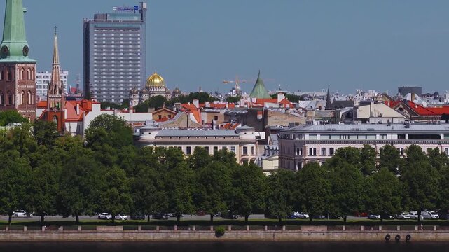 Aerial pan over central Riga, Latvia, with Daugava embankment, parked cars, red tiled roofs, Riga Dome Cathedral clock tower, Nativity Cathedral cupola, and cruise boat moving.
