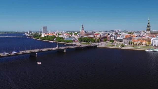 Aerial 4K view of Riga, Latvia, shows Daugava River, Doma Cathedral, St Peters Church, a busy riverside bridge, a tour boat, a moored vessel, and modern towers at midday.