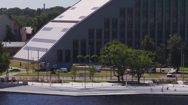 Aerial Riga scene shows National Library of Latvia by Daugava River. Midday sun highlights angled roof and clerestory. People sit by water as cars, a cyclist, and pedestrians move.