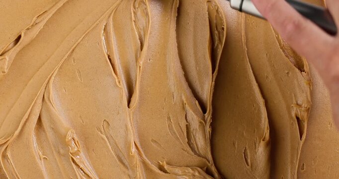 Creamy peanut butter texture background. Top view of a spoon spreading smooth peanut butter across the full frame, creating flowing lines and organic patterns. Macro food texture background.