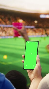 Vertical video: Close-up of mockup phone used by fan in stadium to compare odds during soccer match day, with football supporter checking predictions on live score app on green screen smartphone.