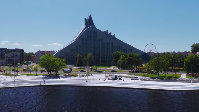 Aerial view shows the National Library of Latvia by the Daugava in Riga, a Ferris wheel beyond trees, cars and vans moving, pedestrians present, midday summer light.