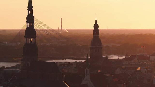 Aerial view over Riga, Latvia at sunset. St Peters Church and Riga Dome Cathedral stand against the Daugava River and Vansu Bridge. Boats move, smoke drifts.