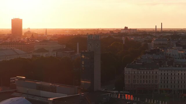 Aerial sunset over central Riga, Latvia, shows the RIGA clock tower with twin faces, tree lined park, church spires, and Opera Hotel as the camera slowly glides.