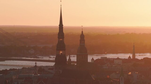 Aerial view of Riga, Latvia at sunset as the drone pans, showing St. Peter's Church and Riga Cathedral above red roofs, the Daugava River, and the Vansu Bridge.
