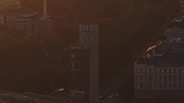 Aerial drone pans and descends above Riga, Latvia, centering a tall clock tower with RIGA lettering and twin clocks, Opera Hotel to the right, park and street below.