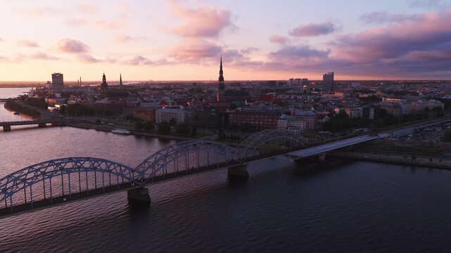 Aerial 4K pan forward over Daugava River in Riga, Latvia shows Railway Bridge, Old Town skyline with St Peters spire, red brick roofs, high rises, and rail lines at sunset