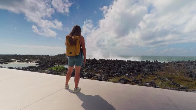 Young Female Traveler Watching Massive Ocean Waves Crash Against Rugged Coastal Rocks Under a Dramatic Sky in Slow Motion