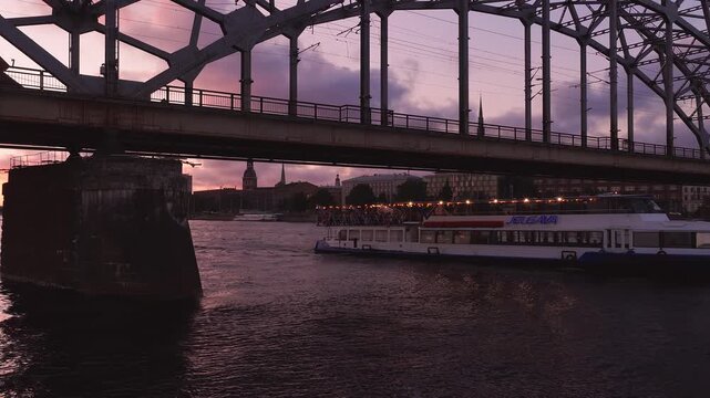 Aerial view shows a riveted steel railway bridge in Riga, Latvia, the Dome Cathedral spires, and cruise boat JELGAVA gliding under, ripples shimmer at twilight.