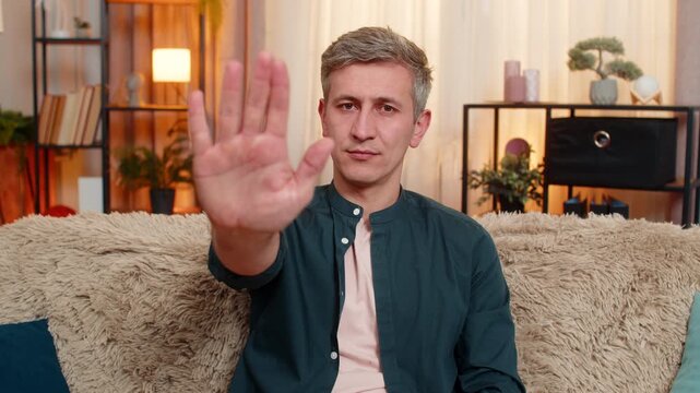 Young man at home raises palm with stop gesture and serious face. Caucasian guy on sofa expresses clear rejection, creating message of personal limit, warning or emotional boundary with strict tone