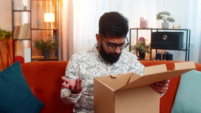 Indian man sitting at home on sofa, opening cardboard box from online delivery and discovering broken wireless headphones. Arabian guy feeling upset and disappointed with damaged product received.