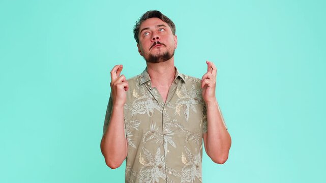 Middle-aged man crossing fingers with tense expression, silently hoping for positive result or good outcome. Young adult guy shows nervous anticipation and emotional hope isolated on blue background.