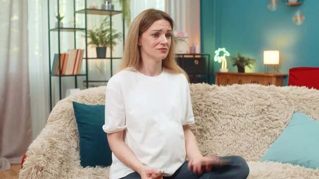 Pregnant middle-aged woman at home watches bitcoin chart fall on smartphone presses phone against forehead sighs worried. Caucasian girl on sofa plans finance strategy saving family money future baby.