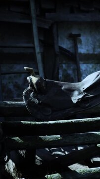 A worn saddle sits on a wooden bench inside a barn. Sunlight filters through the structure, illuminating the saddles faded leather. Tools and equipment are barely visible in the background.
