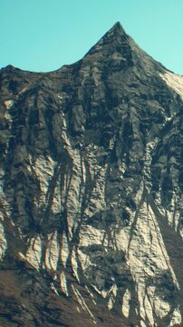 A tall mountain peak stands prominently against the blue sky. The surface shows rocks and snow, with shadows creating patterns in the sunlight. This scene captures the natural landscape.