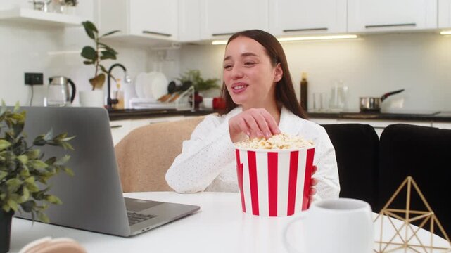 Young woman at home watches trailer on laptop eats popcorn laughs and relaxes fun break. Caucasian girl on sofa sits at table watches video smiles bright mood evening snack casual chill online clip.