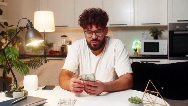 Arabian man at home counts dollar money cash uses calculator checks bills marks paid tracks saving. Egyptian guy at table sorts piles for spend and save plans budget adds note for month late extra.