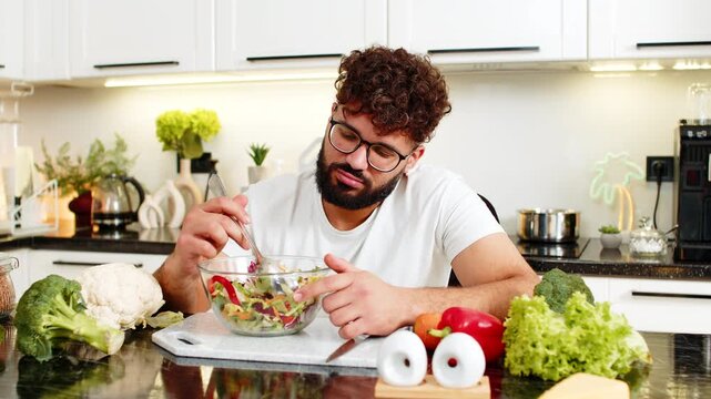 Arabian man forces salad bites in kitchen feeling tired of strict keto diet and healthy nutrition at home. Young guy bored with a diet routine craving comfort food while trying weight loss lifestyle.