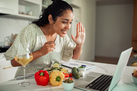 Smiling woman waving to laptop during online cooking class in home kitchen