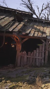 A wooden building stands with damage to its roof in a rural setting. A broken fence surrounds the area. The surroundings show signs of decay with little greenery visible.