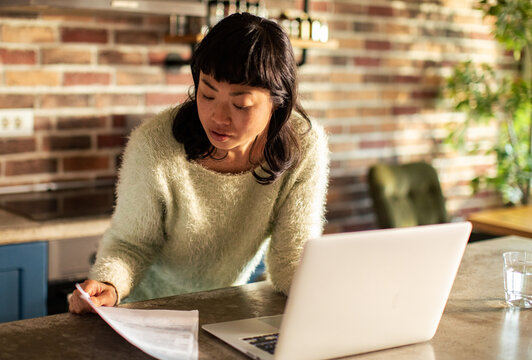Woman reviewing bills at kitchen counter with laptop