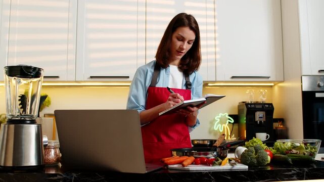 Young woman smiling while studying recipe on laptop writing notes for healthy cooking lesson in kitchen at home. Adult girl planning diet meal prep for nutrition goals indoors during self education.