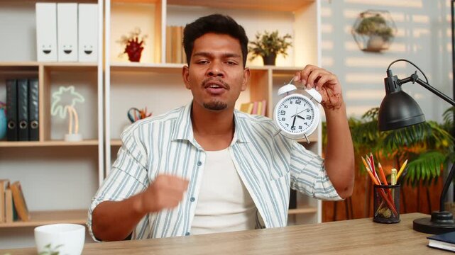 Indian man on home table shows clock to camera and talks with tense face about time during remote work task. Arabian guy on home table explains delay and asks about schedule in calm home office mood.