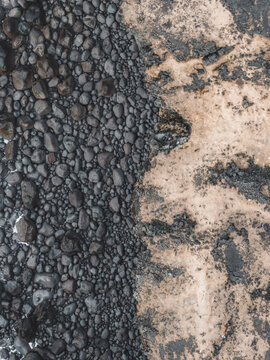Aerial view of black volcanic pebbles contrasting with a textured sandy shore in Iceland.
