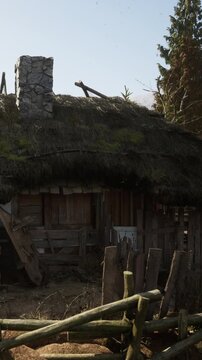 A wooden house with a thatched roof stands in a rural area. The scene shows some trees around the house and a wooden fence nearby. The atmosphere looks simple and rustic in daylight.