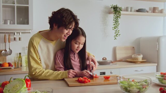 Smiling Young Asian Girl And Male Mentor Collaborating On Cooking In A Bright, Modern Kitchen. Engaged In Food Preparation And Skill Building, Fostering Learning In A Culinary Setting.