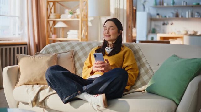 Serene lady using smartphone sitting cozy couch. Woman scrolling cellphone