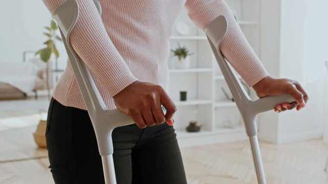 Worried Unrecognizable Black Female Using Crutches For Support While Standing In Modern Living Room, Aiding Recovery At Home. Focus On Rehabilitation And Adaptive Mobility Concept.