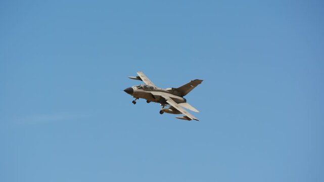 Combat jet in level flight with landing gear extended, side view showing tandem two seat cockpit, external fuel tank under swept wing, military aircraft against clear sky, slow motion
