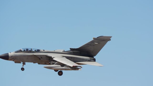 Two seat military jet on final approach, pilots visible under the tandem canopy, landing gear down and external fuel tank under the wing, side view in blue sky, slow motion
