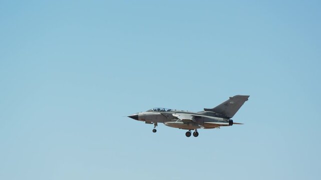 Military jet approaching the runway with landing gear extended, distant side view of combat aircraft flying in vast clear blue sky, external fuel tank visible, slow motion