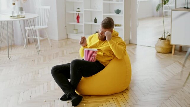 Relaxed Happy Bearded Caucasian Man Enjoying Popcorn While Sitting Comfortably On Yellow Bean Bag In Bright Living Room. Leisurely Moment At Home, Weight Loss Concept.