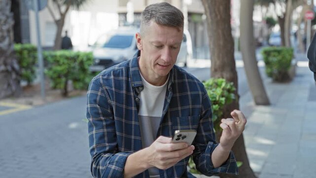 Man holding smartphone with raised fist on a city street bench, reacting with a small victorious smile while checking the screen; excitement personal victory.