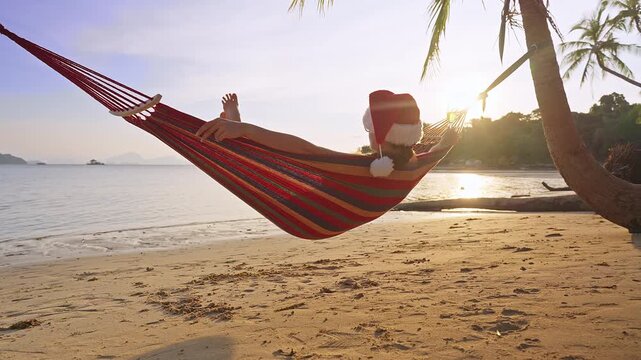Slow Motion Rear View of a Woman Wearing a Santa Hat Relaxing in a Hammock With Sparkling Wine on a Tropical Beach at Sunset, Christmas Vacation, Peace and Freedom