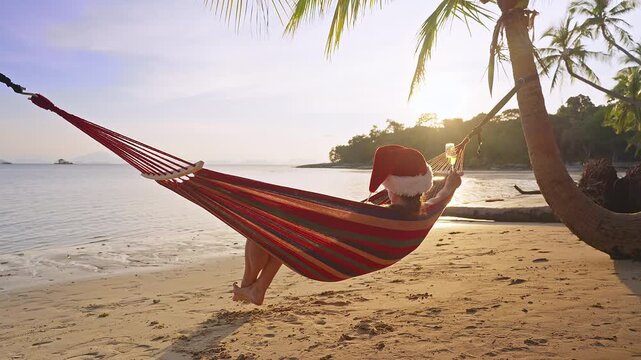 Slow Motion Woman Wearing a Santa Hat Resting in a Hammock While Holding Sparkling Wine Above a Tropical Beach at Sunset, Festive Holiday Relaxation