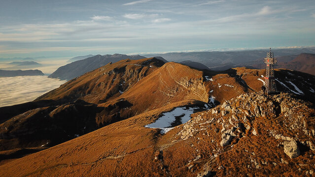 Aerial view of Le Reculet peak with its metal cross, rugged mountain ridges, patches of snow, and a sea of clouds in the valley in Thoiry, Auvergne-Rhone-Alpes, France.