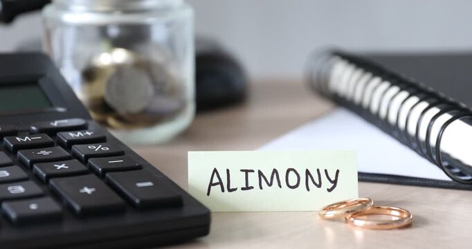 Card with word Alimony, calculator, wedding rings, notebook and coins on wooden background, closeup. Camera zooming in