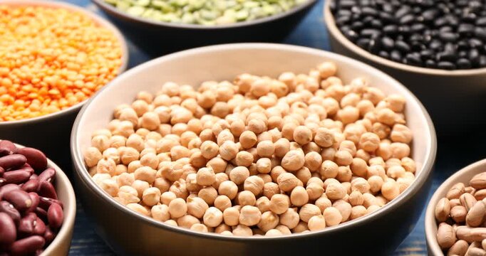 Pouring chickpeas from scoop into bowl, different types of legumes on blue wooden table, closeup