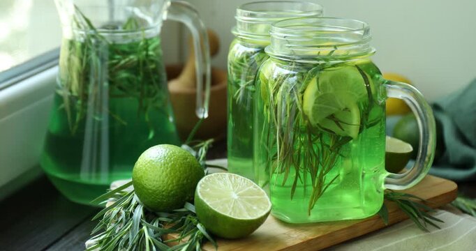 Tasty lemonade with tarragon in mason jars, jug, limes and fresh leaves on wooden table near window, closeup