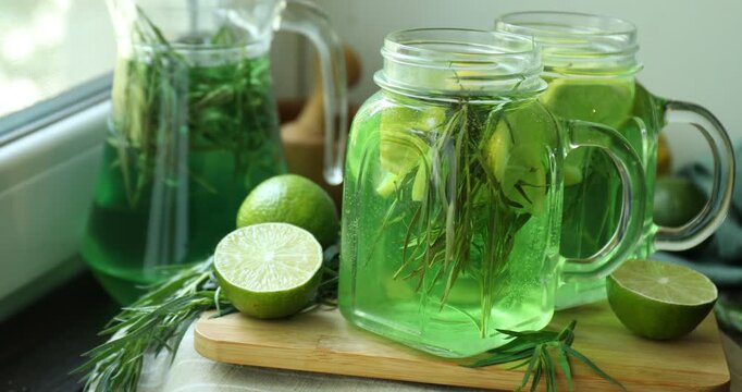 Tasty lemonade with tarragon in mason jars, jug, limes, fresh leaves, mortar and pestle on wooden table near window, closeup
