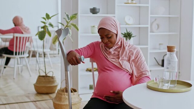 Worried Pregnant African American Woman Sitting At Home, Monitoring Baby's Health With Device, Holding Her Belly, In Comfortable Indoor Setting. Focus On Motherhood And Healthcare Concept.