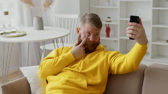 Smiling Healthy Bearded Man In Yellow Hoodie Taking Fun Selfie With Smartphone While Sitting Comfortably At Home. Casual Atmosphere In Modern Living Room. Lifestyle Concept.