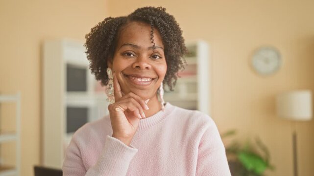 Young woman hand to chin, smiling and wearing earrings with pink sweater in studio, relaxed pose and direct eye contact; thoughtful reflection.